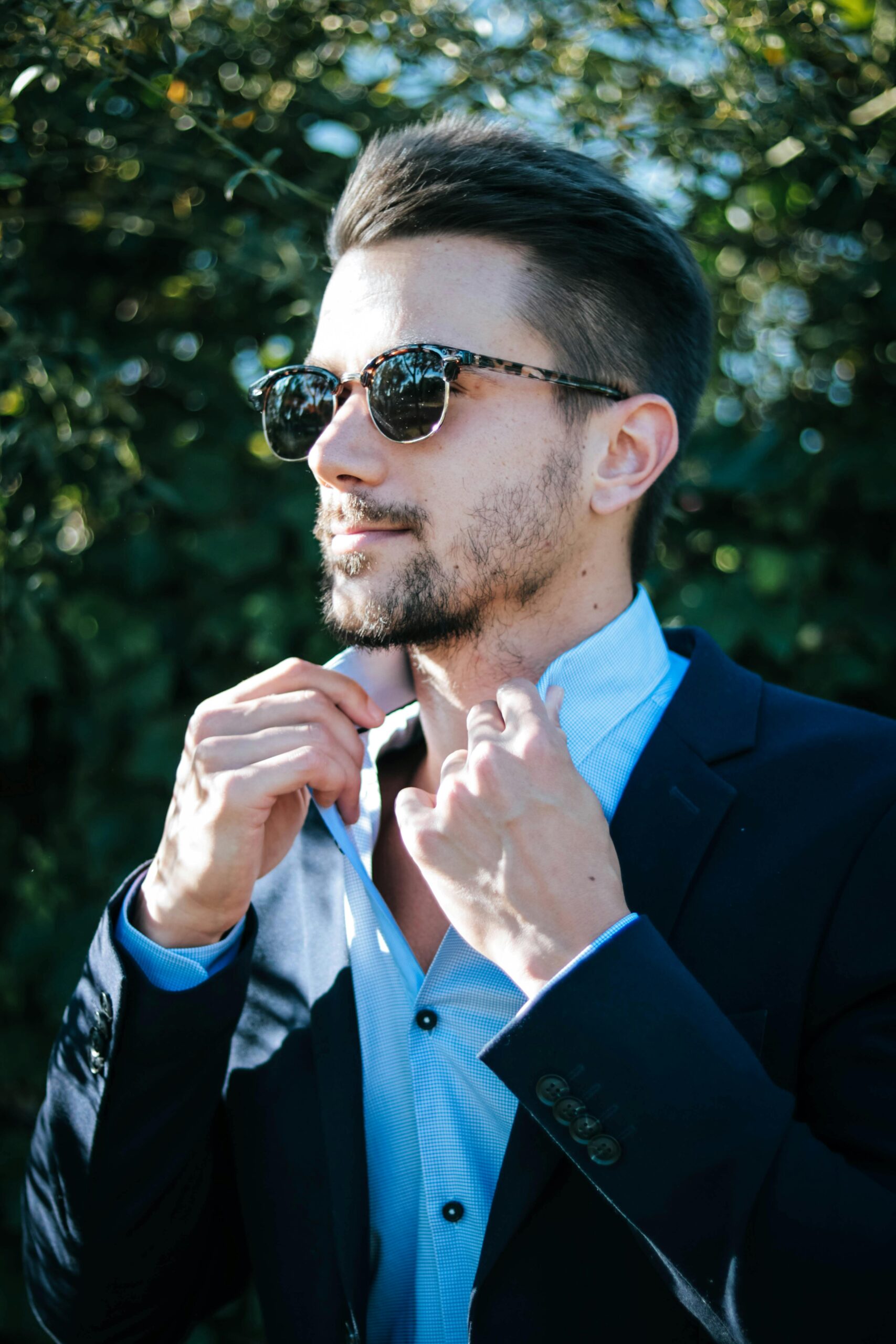 A man in sunglasses adjusts his shirt collar while wearing a blue suit jacket. He is outdoors with greenery in the background.