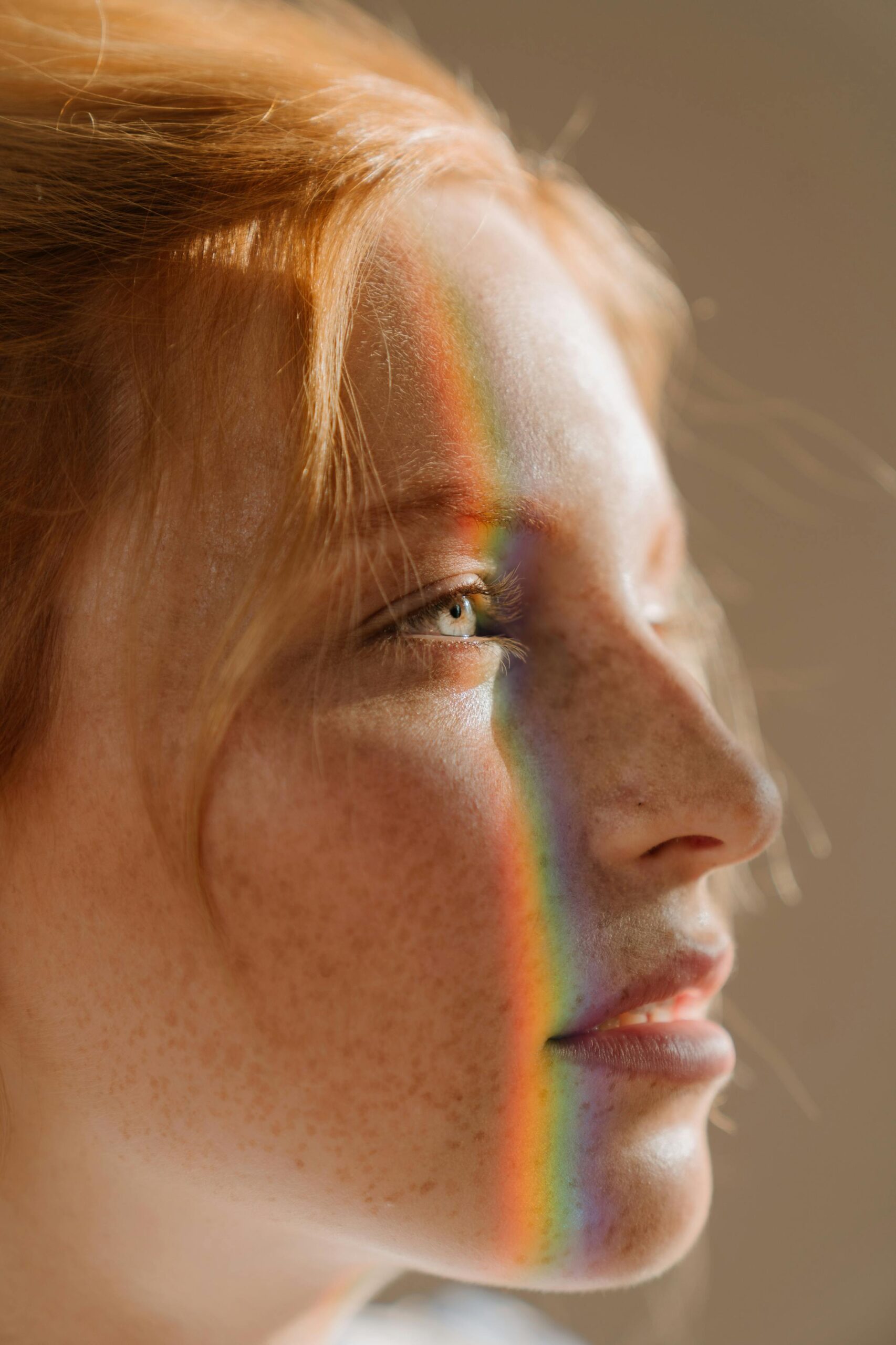 Close-up of a person's face with freckles and red hair, illuminated by a rainbow light reflection across their eyes and nose.